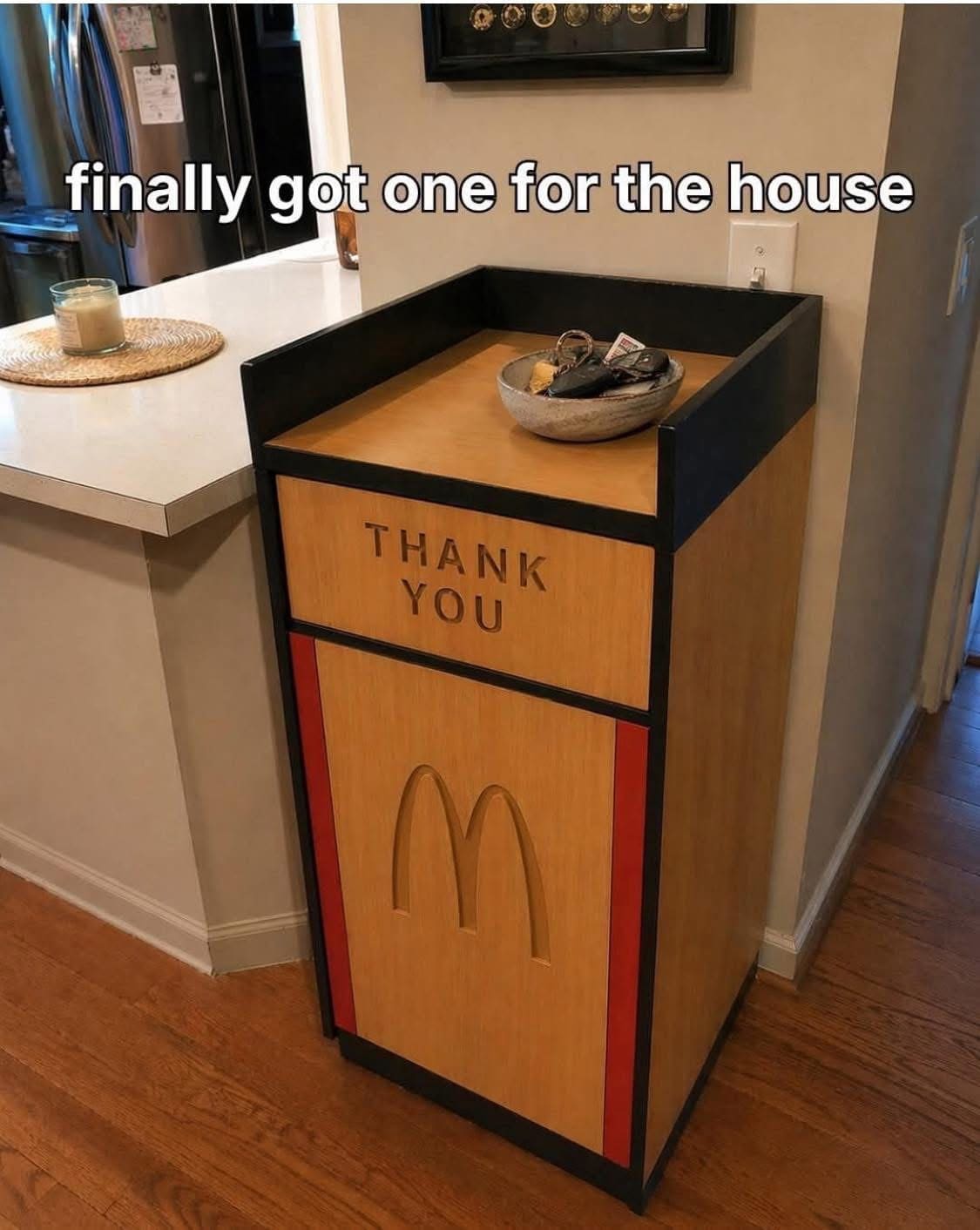 A domestic meme dump showing a classic wooden McDonald's "THANK YOU" trash bin and tray return station repurposed as furniture in a home's entryway. It is currently holding a bowl of car keys.