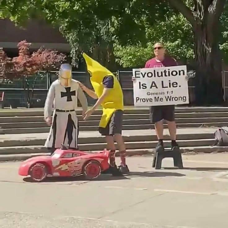 A surreal photo from a meme dump capturing a chaotic public encounter. A man in full crusader knight armor stands next to a person in a banana costume who is gently placing a toy Lightning McQueen car on the pavement. In the background, a man stands on a step-stool holding a sign that reads, "Evolution Is A Lie. Genesis 1:1 Prove Me Wrong."