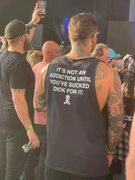 A high-octane funny meme captured in a crowd. A man’s back is visible, sporting a black tank top with bold white text that declares: "IT’S NOT AN ADDICTION UNTIL YOU’VE SUCKED DICK FOR IT."