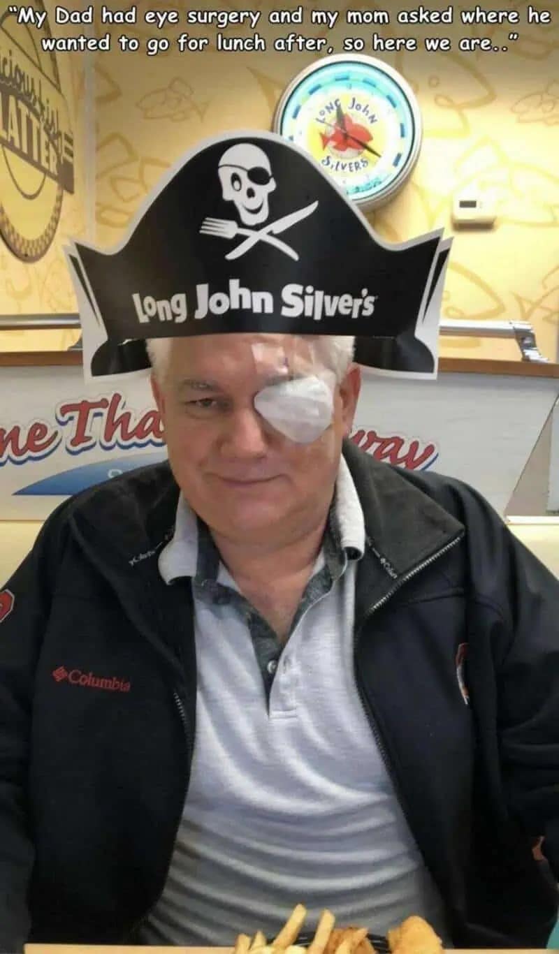 wholesome and funny meme of an older man sitting in a Long John Silver's restaurant. He is wearing a paper pirate hat that perfectly aligns with a large white medical patch over his eye from recent surgery, making him look like a literal pirate while he eats his fish and chips.