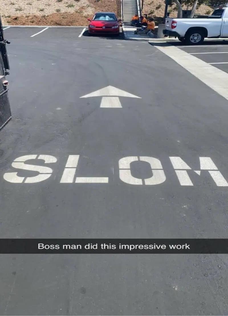 A classic "you had one job" funny meme showing a freshly paved road. Large white stencils intended to warn drivers to go "SLOW" have been misspelled in giant letters as "SLOM," complete with a white arrow pointing forward.