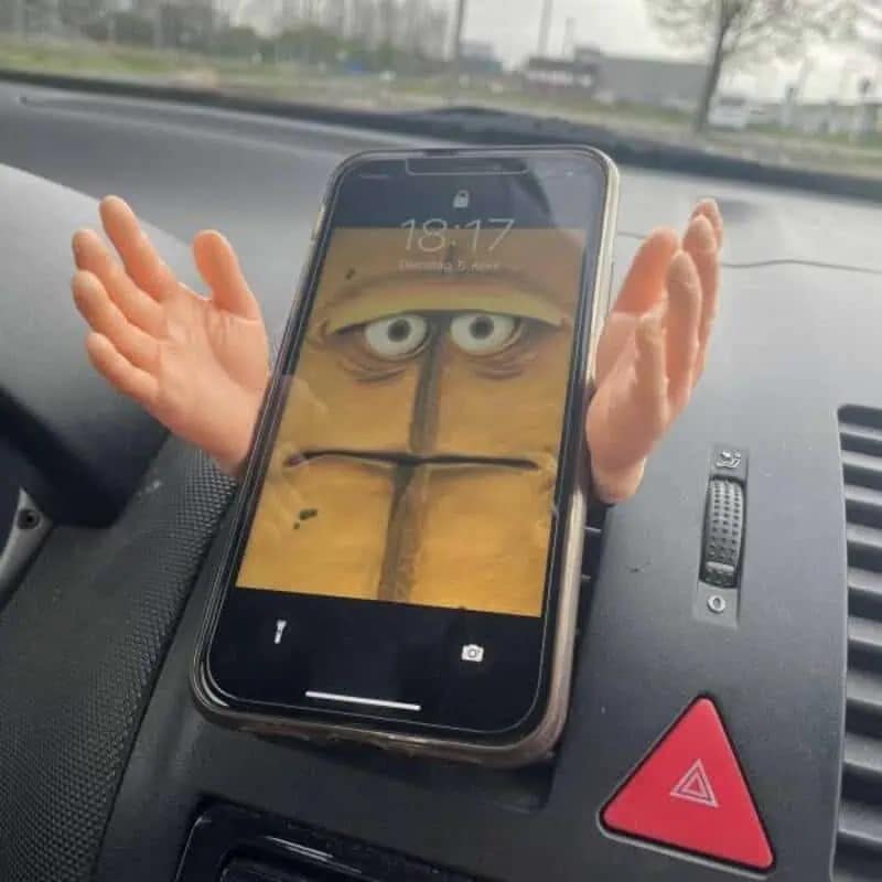 creative funny meme featuring a smartphone mounted on a car dashboard. The phone holder is shaped like two tiny, realistic human hands that appear to be throwing themselves up in exasperation, perfectly matching the tired, annoyed face set as the phone's wallpaper