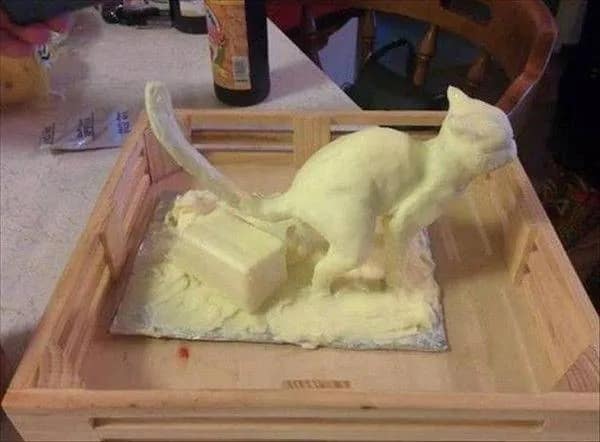 A photo of a rustic wooden tray containing a large block of yellow butter. Standing on top is a crude but unmistakable sculpture of a cat, also made of butter, posed as if it is using the block beneath it as a litter box.