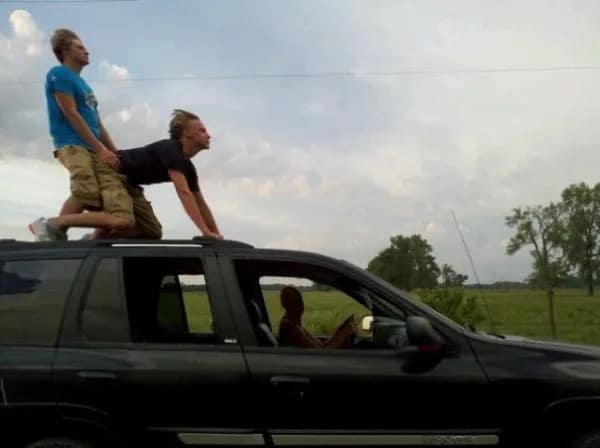 A chaotic and hilarious meme showing two men on the roof of a moving black SUV. One man is on all fours mimicking an animal while the other sits behind him, both looking ahead with serious expressions as the car drives past a rural field.