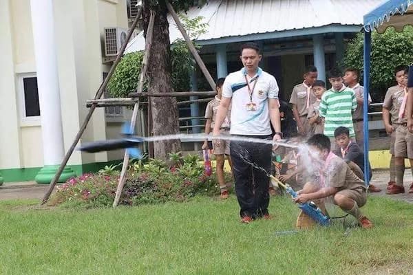 A high-chaos funny meme capturing a science demonstration gone wrong. While a teacher and students watch a water rocket launch, the bottle stays grounded, blasting a direct, high-pressure stream of water directly into the face of a crouching boy scout.