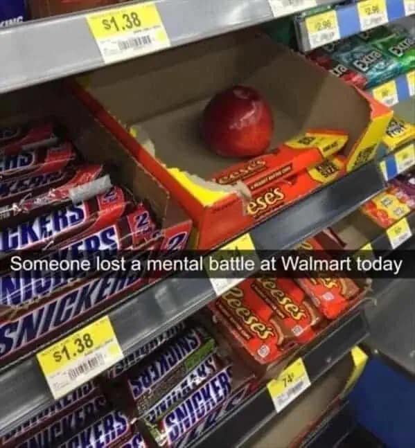 A relatable funny meme from a Walmart aisle showing a single, healthy red apple abandoned in a cardboard display box filled with Reese’s Peanut Butter Cups and Snickers bars. The Snapchat-style caption reads: "Someone lost a mental battle at Walmart today."