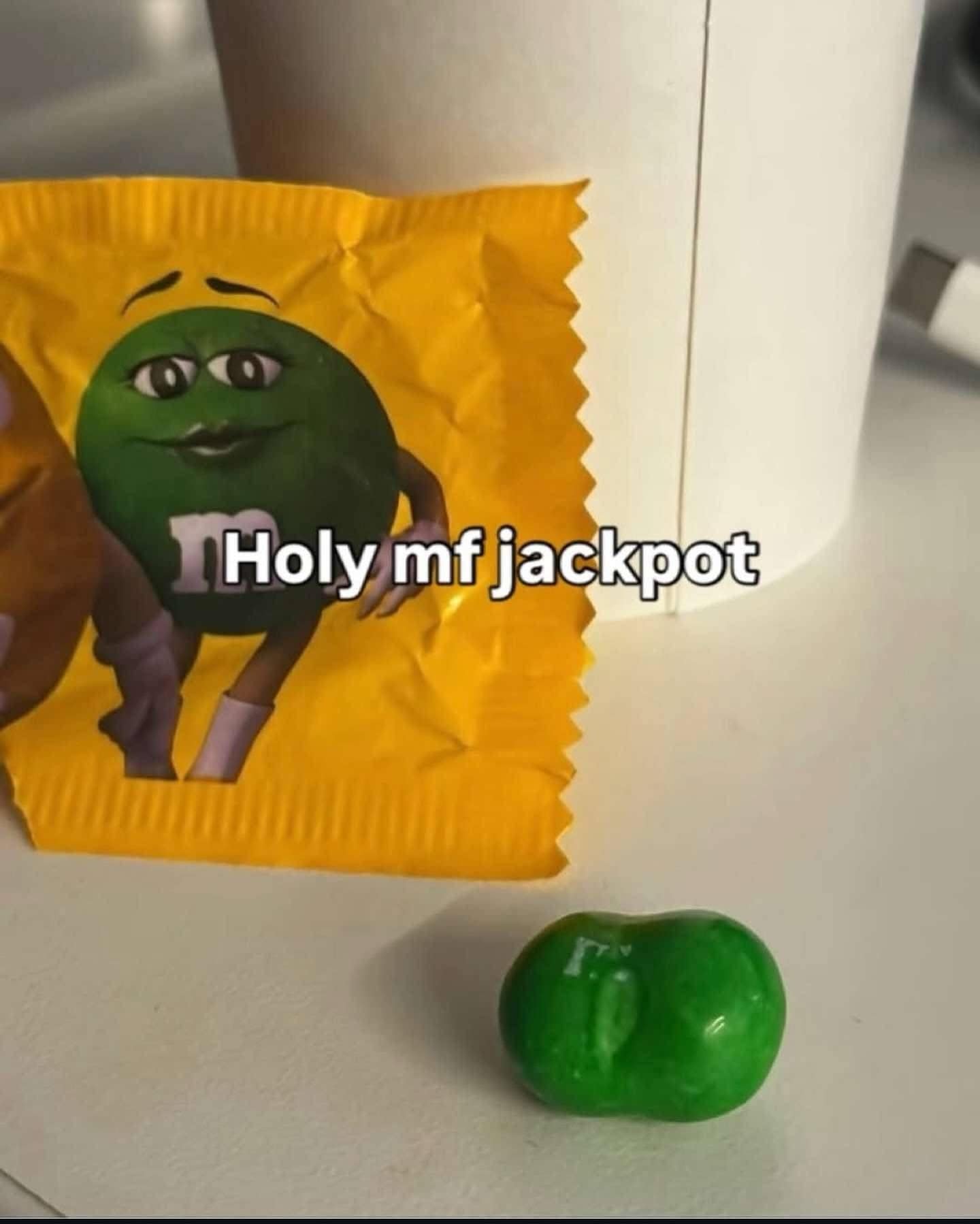 A lucky find in a meme dump featuring a single green M&M candy that is uniquely misshapen, appearing as two candies fused together to form a distinct "butt" shape. The text overlay reads, "Holy mf jackpot."