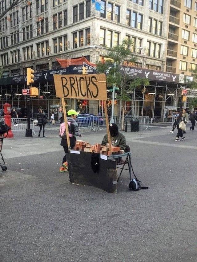 An urban meme dump snapshot taken in a busy city square. A person in a black balaclava sits behind a cardboard sign that simply reads "BRICKS," with a pile of actual red bricks stacked on a table in front of them.
