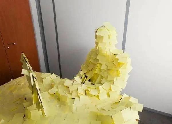 A visual metaphor for crippling anxiety or a massive to-do list: a person sitting at a desk is entirely covered from head to toe in layers of yellow sticky notes, with their laptop and workspace similarly buried in paper reminders.