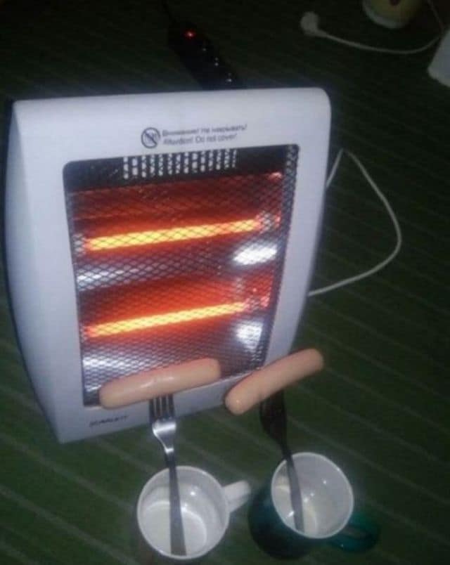 A low-budget culinary funny meme. Two sausages are skewered onto forks and positioned inches away from the glowing red coils of an electric space heater. Two white mugs sit below to catch any drippings.