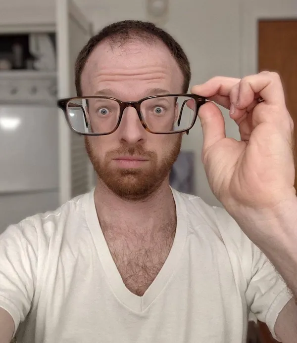 A disorienting meme dump selfie of a man holding a pair of extremely thick-lensed glasses in front of his face. The high-prescription lenses create a massive, wide-eyed distortion that makes his eyes appear huge and cartoonish compared to his actual head size.