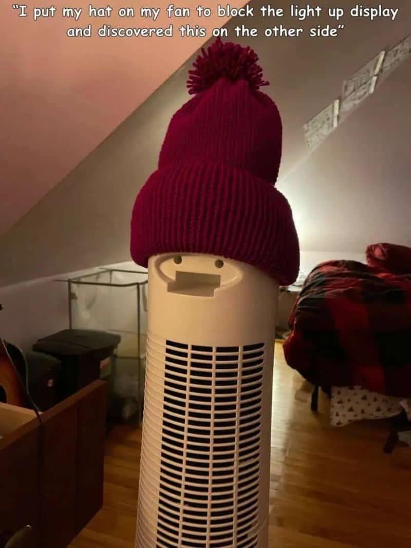 A classic "accidental face" hilarious meme. A tall white tower fan has a red pom-pom beanie pulled over the top to hide a light-up display. From the back, the fan’s handle and shape create the appearance of a tall, thin character with a wide, gaping mouth wearing a hat.