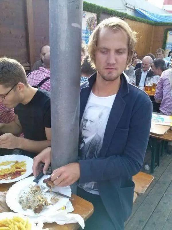 A peak "awkward positioning" hilarious meme from a crowded outdoor beer garden. A man in a blue blazer is squeezed so tightly against a massive grey support pole that he has to wrap his arms around it to eat his fries. Ironically, his t-shirt features a portrait of Sigmund Freud.