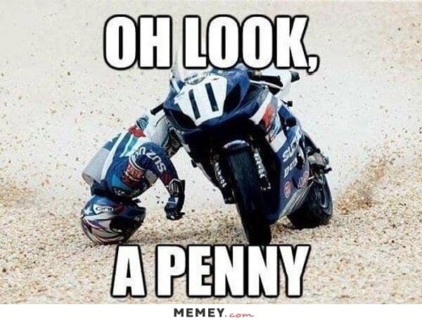 A "look on the bright side" motorcycle meme showing a racer mid-crash, sliding across the gravel trap with their head pinned to the ground. The large white caption turns the disaster into a joke: "OH LOOK, A PENNY."