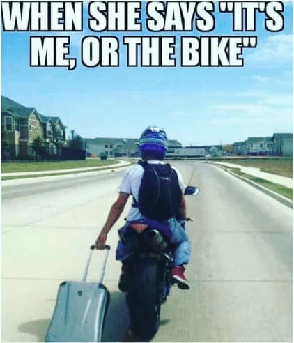 A definitive "commitment" motorcycle meme. A rider in a purple helmet is seen from behind, riding down a residential street while pulling a silver rolling suitcase behind them with one hand. The caption: "WHEN SHE SAYS 'IT'S ME, OR THE BIKE'."