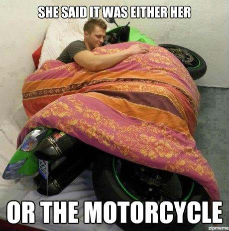 A "love story" motorcycle meme where a man is seen in a bedroom, tucked into bed and sleeping soundly under a pink and orange patterned blanket next to his green sportbike. The text: "SHE SAID IT WAS EITHER HER OR THE MOTORCYCLE."