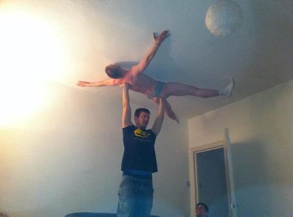 A bizarre no context image of a man in a black t-shirt effortlessly hoisting another man up to touch the ceiling with a powerful one-handed lift, creating a weird human-sized ceiling ornament in a plain, dimly lit room.