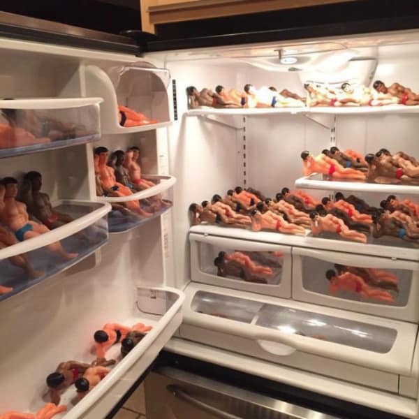 A legendary no context image of an open refrigerator completely packed with dozens of muscular male action figures in swim trunks, all posed lounging across the shelves, egg trays, and door bins in a cold, plastic party.