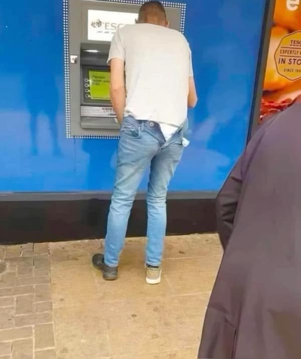A baffling no context image from a parking lot showing a man at an ATM with his light-wash jeans pulled down to his mid-thighs, standing completely unbothered while conducting his transaction in a white t-shirt.