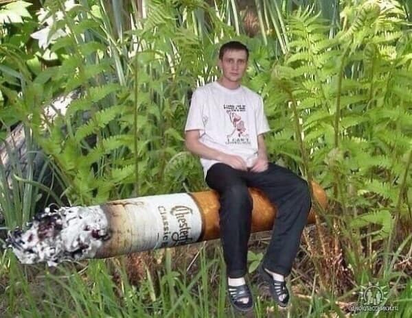 A surreal and grainy cursed image of a young man in socks and sandals sitting cross-legged atop a giant "Chesterfield" brand cigarette that appears to be the size of a log in a dense forest of ferns.