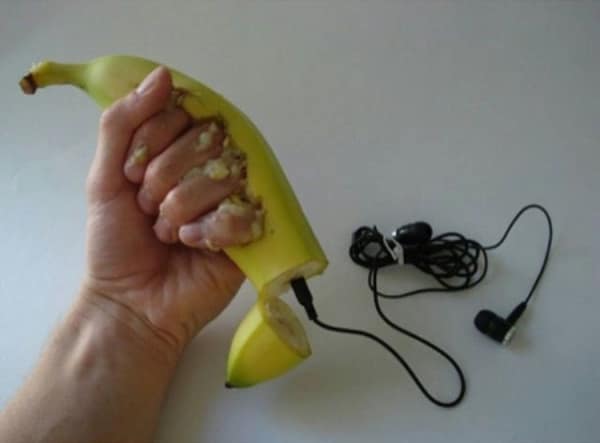 A deeply uncomfortable cursed image where a human hand is shown crushing a banana, with the severed end of the fruit revealing a 3.5mm headphone jack plugged directly into the mushy center.
