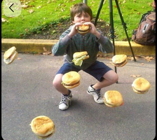 A bizarre no context image featuring a young boy in a defensive crouch eating a cheeseburger while several other burgers dangle around him on thin strings, creating a frozen rain of fast food in the middle of a park.