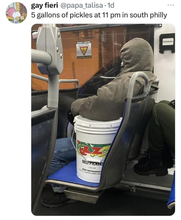 A peak South Philly no context image captured on a bus at night, featuring a person in a heavy parka sitting with a massive, 5-gallon industrial bucket of deli pickles as their only seat-mate.