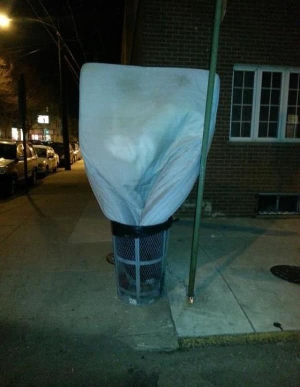 A haunting no context image from a dark city street where someone has managed to stuff a full-sized queen mattress vertically into a standard wire mesh sidewalk trash can, creating a ten-foot-tall white monolith.