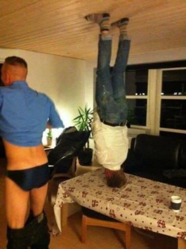A chaotic no context image showing a man balanced perfectly on his head on a dining table with his feet pressed against the wooden ceiling, while a friend in the foreground casually walks by with his pants around his ankles.
