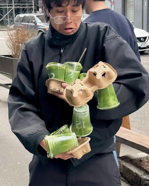 A high-stress no context image capturing the exact millisecond a cardboard drink carrier fails, sending several plastic cups of bright green matcha lattes plummeting toward the pavement as the carrier's owner looks on in horror.