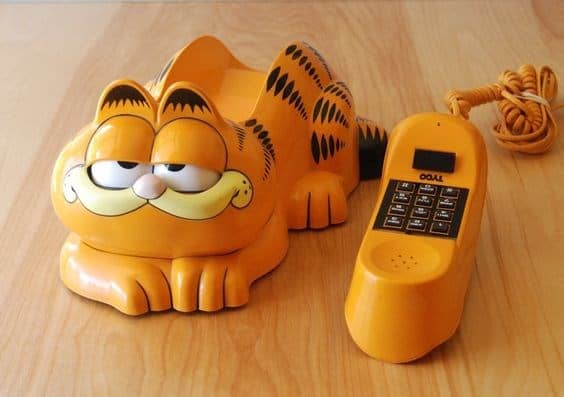 An iconic novelty landline phone shaped like Garfield the cat. The lazy orange tabby's eyes open and close depending on whether the receiver is docked. The matching orange handset features a standard black push-button keypad