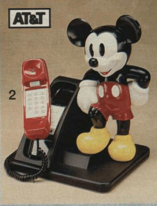 A novelty landline phone starring a classic Mickey Mouse figurine standing on a black pedestal. A red push-button handset sits in a cradle to his left.
