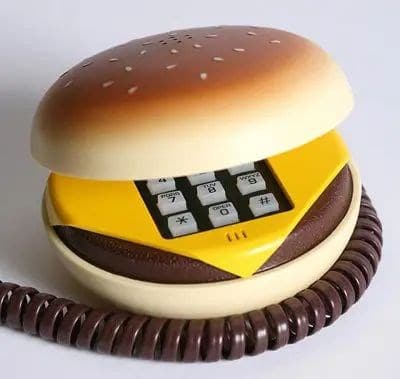 A classic novelty landline phone designed to look like a cheeseburger. The top bun acts as the receiver, lifting away to reveal a numerical keypad nestled between the "cheese" and "patty."