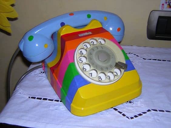 A vibrant vintage landline phone with a rotary dial, custom-painted with a multi-colored rainbow stripe pattern. The light blue handset is decorated with playful green and red dots.
