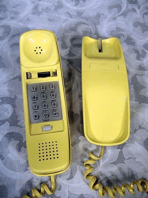 A sleek vintage landline phone in a monochromatic soft yellow. The keypad is integrated into the slim handset, which rests on a matching wall-mountable base.