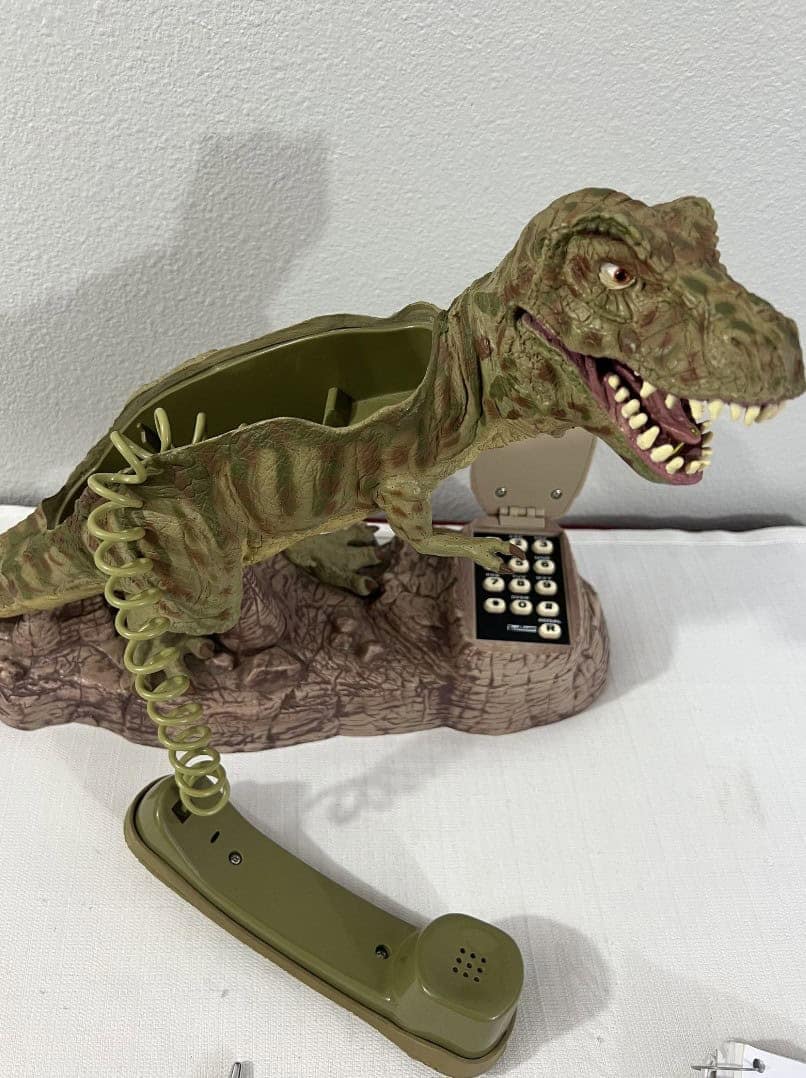fierce novelty landline phone featuring a highly detailed Tyrannosaurus Rex sculpt. The T-Rex stands over a rocky base that holds the keypad, and a classic green handset is attached via a coiled olive-drab cord.