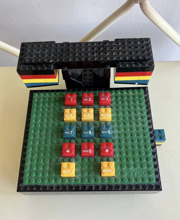 A colorful novelty landline phone constructed from building blocks, made by Tyco. The square base looks like a green LEGO-style plate with primary-colored buttons, while the boxy handset features a matching black, yellow, red, and blue stacked aesthetic.