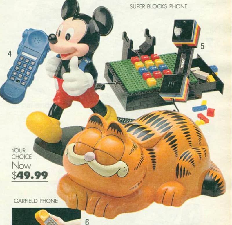 A catalog clipping showing several novelty landline phones available for $49.99. Featured are a Mickey Mouse figure holding a phone, the Tyco Super Blocks phone, and the legendary Garfield cat phone.