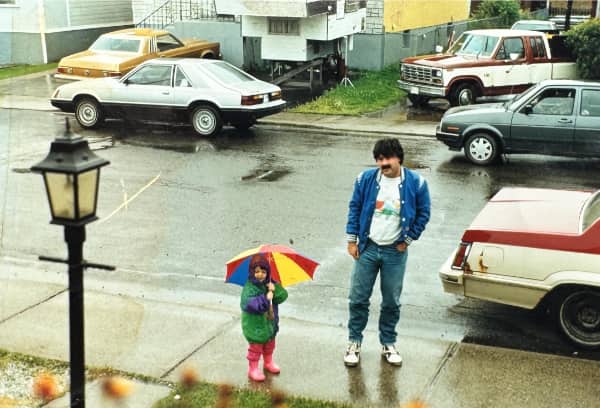 Father and child with colorful umbrella standing in rainy street surrounded by vintage 1980s cars