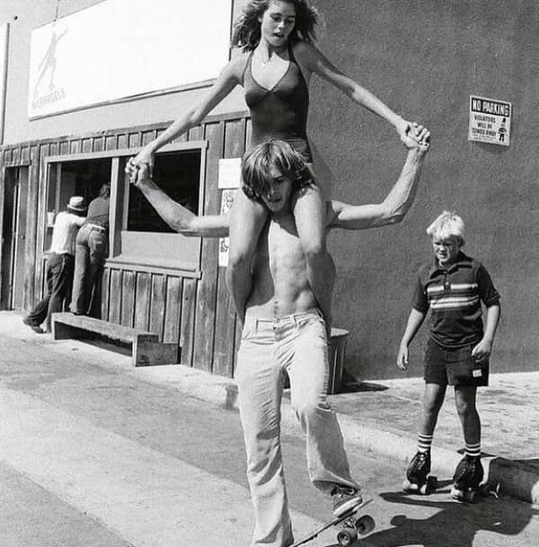 Shirtless 1970s skateboarder carrying girl on shoulders outside Venice Beach-style boardwalk shop