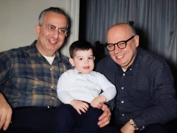 Two middle-aged men with thick-framed glasses sitting on couch with smiling toddler between them
