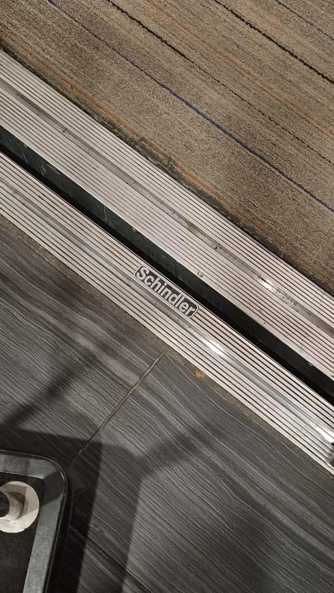 dark punny meme featuring a close-up of a metal floor threshold. Embossed clearly in the center of the metal strip is the manufacturer’s name: "Schindler," making the object a literal "Schindler's List."