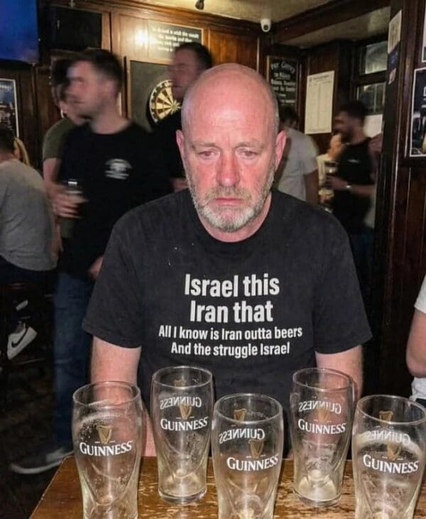 A weary-looking man sits in a pub behind four empty Guinness glasses wearing the ultimate geopolitical punny meme t-shirt. The text reads: "Israel this Iran that. All I know is Iran outta beers and the struggle Israel."