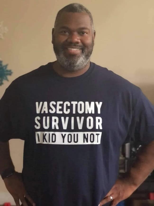 A confident man smiles while wearing a classic medical punny meme shirt. The navy blue t-shirt features bold white letters that read: "VASECTOMY SURVIVOR I KID YOU NOT," referencing the lack of "kidding" as a result of the procedure.