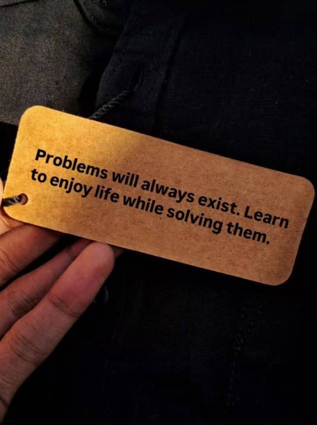 A hand holding a brown cardboard tag with advice about enjoying life while solving problems.