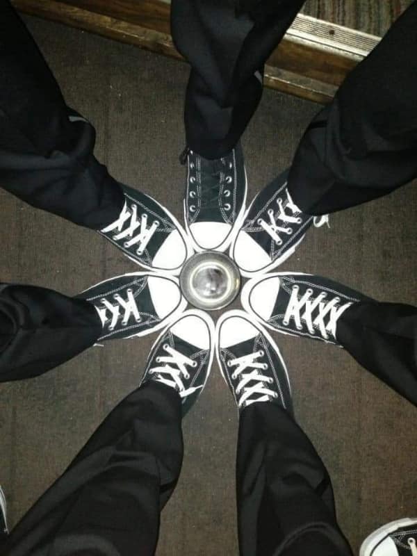 geometrically perfect satisfying photo of seven friends standing in a circle, their identical black Converse sneakers pointed inward toward a central round metal plate, creating a star-like symmetry on the dark carpet.