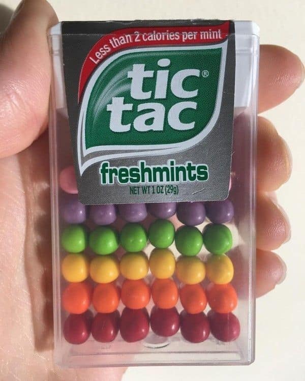 A clear Tic Tac container held in a hand, showcasing a satisfying photo of the mints meticulously sorted into five vibrant, monochromatic layers of purple, green, yellow, orange, and red.