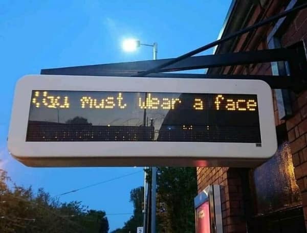 A glitchy public LED sign displaying an accidental horror prompt—a tech fail where the intended "mask" message is cut off, leaving only the ominous command: "You must wear a face."