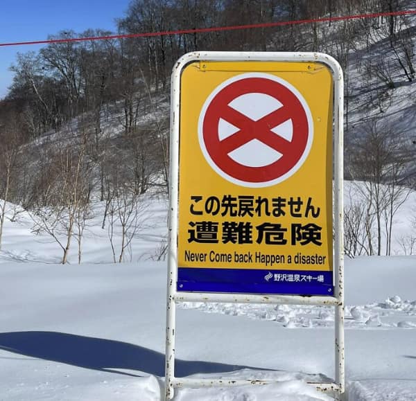 An ominous translation fail on a yellow warning sign in a snowy mountain range. The Japanese warning about the dangers of getting lost is translated into a terrifying English command: "Never Come back Happen a disaster."