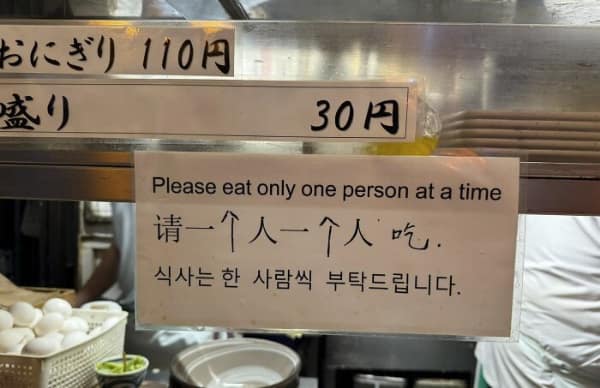 A cannibalistic translation fail at a busy food counter. A sign meant to manage the queue or portion sizes instead tells customers: "Please eat only one person at a time."
